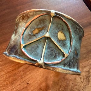 May include: A wide, bronze-colored cuff bracelet with a peace symbol in the center. The peace symbol is made of copper and gold-colored metal. The bracelet has a textured surface with a turquoise patina.