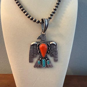 May include: Silver-tone pendant necklace with a bird design. The pendant features an orange, teardrop-shaped stone and two small turquoise stones. The necklace has a beaded chain.