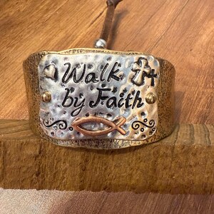 May include: A wide cuff bracelet with a hammered silver-tone metal plate featuring the embossed black text Walk by Faith. The plate includes a small heart, a cross, and a copper-tone fish symbol. The bracelet has a dark bronze-tone finish.