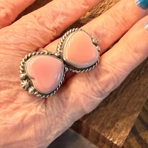 May include: A silver ring featuring two smooth, light pink heart-shaped stones set side-by-side. Each stone is framed by a twisted rope-style metal border with small silver bead accents at the edges, creating a symmetrical, elongated design.