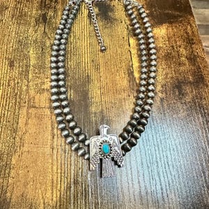May include: A double-strand necklace featuring round, metallic silver-toned beads and a central pendant shaped like a bird with etched geometric patterns and a single, oval-shaped turquoise-colored stone set in the center of the bird's chest.