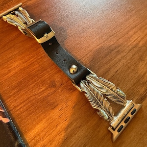 Op de afbeelding: Een zwarte leren horlogeband met goudkleurige veermotieven en een zilveren gesp. De band heeft een decoratief ontwerp met twee grote veren en kleine turkooizen stenen.