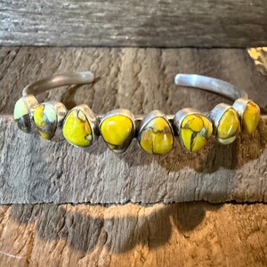 May include: A silver cuff bracelet featuring eight teardrop-shaped yellow stones with dark brown matrix veining. The stones are set in individual silver bezels along the front of the polished metal band, which has an open back design.
