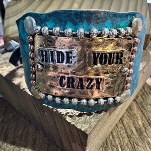 Puede incluir: Una pulsera de puño turquesa y cobre con las palabras "HIDE YOUR CRAZY" en letras plateadas y cobrizas. La pulsera está decorada con cuentas y remaches plateados y cobrizos.