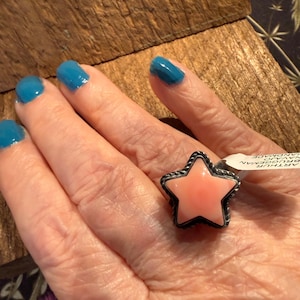 May include: A silver ring featuring a star-shaped, smooth, light pink stone set in a twisted metal border. A small white tag attached to the ring band displays the text ARTHUR BRUGGEMAN NAVAJO HANDMADE in black ink on a white background.