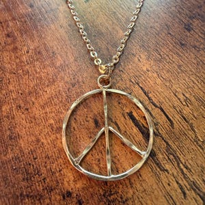 May include: Gold-coloured peace sign pendant necklace. The pendant is a circular design with a vertical line and two diagonal lines, representing peace. The necklace chain is also gold-coloured and hangs on a wooden surface.