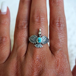 May include: A silver ring featuring a bird design with spread wings and a small, oval-shaped turquoise stone set in the centre of the body. The metal has a textured, oxidised finish that highlights the detailed feather patterns on the wings.
