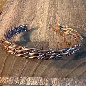 May include: A copper-toned cuff bracelet featuring a multi-strand design with alternating smooth, thick wires and thinner, twisted rope-like wires woven together in a continuous pattern, forming an open-ended circular band with rounded ends.
