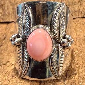 May include: A wide silver ring featuring a central oval-shaped smooth pink stone. The band is decorated with four embossed leaf motifs, two on each side of the stone, and small clusters of silver spheres positioned between the leaves on both sides.