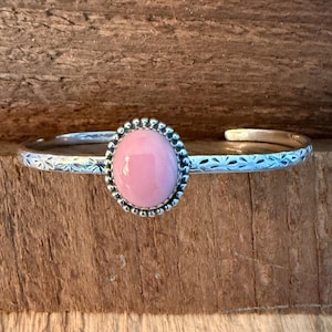 May include: A silver cuff bracelet with a large, oval, light pink stone set in a beaded bezel. The band features a repeating starburst pattern. The bracelet is displayed on a wooden surface.