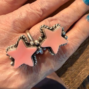 Pode incluir: Um anel de prata com duas incrustações em forma de estrela de pedra lisa cor-de-rosa clara. Cada estrela é emoldurada por um rebordo de fio de prata torcido e acentuada com pequenas contas de prata. O aro do anel é liso e polido.