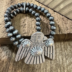 May include: A double-strand bracelet featuring round, metallic grey beads and a central silver-toned pendant shaped like a stylised bird. The bird pendant displays etched geometric patterns, radiating lines on the wings, and a circular centre detail.