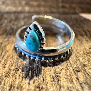 May include: A silver ring featuring a teardrop-shaped turquoise stone. The ring has a detailed band with a beaded design and a decorative setting around the stone. The turquoise stone has brown and blue hues.