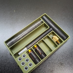 Puede incluir: Una caja de plástico verde con compartimentos para guardar objetos pequeños. La caja contiene una herramienta en forma de T negra, tres proyectiles con punta amarilla, un pequeño tubo de objetos negros y ocho objetos circulares negros.