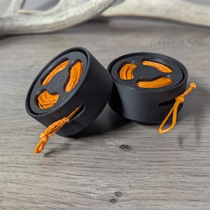 Op de afbeelding: Twee zwarte, cilindrische containers met oranje touw aan de binnenkant en een lus van oranje touw die vanaf de zijkant uitsteekt. De containers staan op een grijs houten oppervlak, met een vage geweistak op de achtergrond.