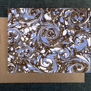 May include: A greeting card with a marbled design in shades of brown, blue, and white. The card is rectangular and has a swirling pattern. A brown envelope is beneath the card.