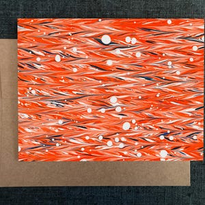 May include: A greeting card with a marbled design in orange, white, and navy blue, with a kraft paper envelope. The card's pattern features flowing lines and white circular accents, creating a textured appearance. The card and envelope are set against a dark gray background.