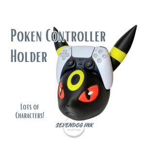 May include: A black and yellow character-themed controller holder with a white controller. The holder has red eyes and a yellow circle. Text on the holder reads "Poken Controller Holder" and "Lots of Characters!"