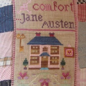 May include: A cross-stitched wall hanging with the quote "There's nothing like staying home for real comfort" by Jane Austen. The design includes a house, trees, and floral elements, all in shades of pink, brown, and blue.