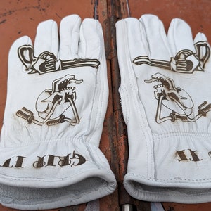 May include: A pair of white leather work gloves with brown etched designs. The gloves feature a key and wrench design, with the words "GRIP IT" etched on the cuff.