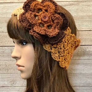 May include: A crocheted headband with a large brown flower and a brown button. The headband is made in shades of brown, tan, and cream.