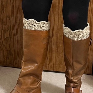 May include: A pair of brown leather knee-high boots with a buckle detail. The boots are worn with black leggings and beige crocheted boot cuffs. The boots have a slight shine and a classic design.