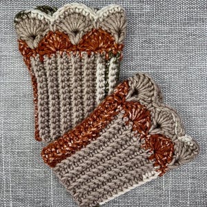 May include: Two handmade crochet arm warmers in shades of brown, tan, and rust. The arm warmers feature a ribbed design with a scalloped edge. The yarn appears to be a soft, textured material, perfect for cooler weather. The arm warmers are displayed on a textured gray surface.