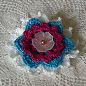 May include: A crocheted flower with a pink center and blue petals, accented with a clear plastic button with a gold center.