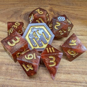 May include: A set of polyhedral dice for tabletop games. The dice are a translucent reddish-brown with gold-colored numbers. A central hexagonal piece features a gold border and a white and gray graphic.