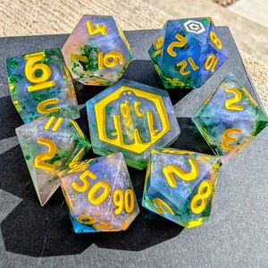 May include: A set of seven polyhedral dice with a fantasy theme. The dice are clear with blue, green, and yellow accents. The numbers are yellow. The dice are arranged on a dark surface.