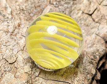 May include: A yellow glass marble with white swirls. The marble has a clear center and is surrounded by yellow swirls.