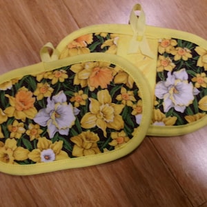 May include: Two yellow and floral patterned oven mitts. The mitts have a black background with yellow, white, and orange daffodils. The edges are trimmed with yellow fabric and have a loop for hanging.