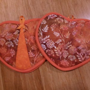 May include: Two heart-shaped pot holders with a floral pattern in shades of orange, red, and white. Each pot holder has a loop for hanging and a solid orange accent. The edges are trimmed with orange fabric.