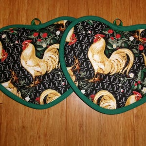 May include: Two heart-shaped pot holders with a black background featuring a rooster design. The roosters are beige and red, with green trim and hanging loops. The design includes red berries and green foliage.