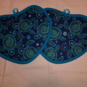May include: Two navy blue, heart-shaped pot holders with a teal turtle pattern and a teal border. Each pot holder has a small loop for hanging.