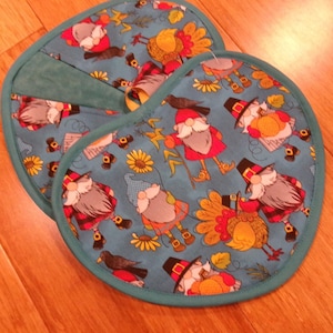 May include: Two turquoise pot holders with a Thanksgiving theme. The fabric features cartoon gnomes, turkeys, sunflowers, and crows. The pot holders have a teal green trim and a loop for hanging.