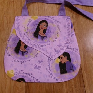 May include: A small, lavender fabric purse with a flap closure and a long strap. The purse is decorated with illustrations of a woman with long black hair, surrounded by floral and butterfly designs. The background is a light purple color.