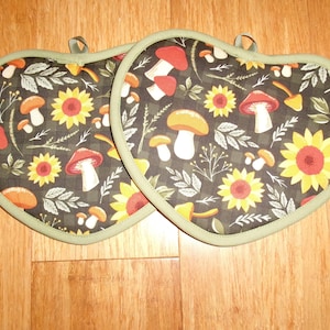 May include: Two heart-shaped pot holders with a dark background featuring sunflowers, mushrooms, and foliage. The pot holders have a green trim and a loop for hanging. The design includes red, yellow, and white elements.
