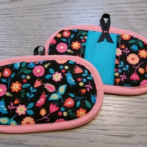 May include: Two pink and black oven mitts with a floral and bird pattern. The mitts have a turquoise lining and a black ribbon.
