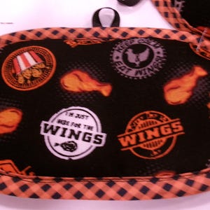K&ouml;nnte beinhalten: Schwarzer und orangefarbener Topflappen mit H&auml;hnchenfl&uuml;gel-Motiven und dem Text "I'm just here for the wings". Der Topflappen hat eine schwarze Schlaufe zum Aufh&auml;ngen und einen karierten Rand.