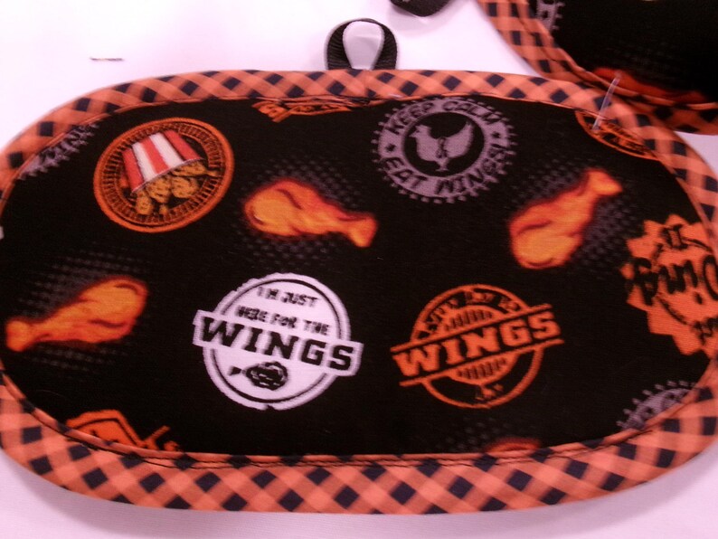 K&ouml;nnte beinhalten: Schwarzer und orangefarbener Topflappen mit H&auml;hnchenfl&uuml;gel-Motiven und dem Text "I'm just here for the wings". Der Topflappen hat eine schwarze Schlaufe zum Aufh&auml;ngen und einen karierten Rand.