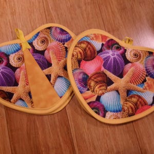 May include: Two heart-shaped potholders with a vibrant seashell pattern. The design features starfish, seashells, and sea urchins in shades of pink, blue, purple, and brown, set against a yellow border. Each potholder has a yellow loop.