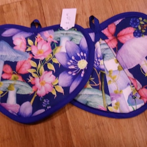 May include: Two heart-shaped pot holders with a dark blue background. The pot holders feature a floral design with pink flowers, purple mushrooms, and green leaves. The edges are trimmed with a dark blue binding.