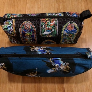 May include: Two zippered pencil cases with different designs. The top case is black with a stained glass pattern featuring a video game character. The bottom case is blue with a video game character design.