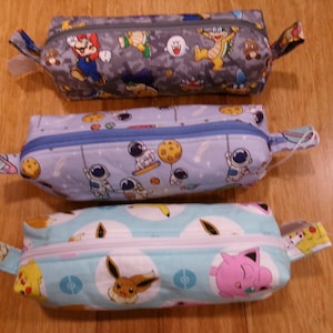 May include: Three zippered pencil cases with different colorful patterns. The first case has a gray background with a Mario and Luigi theme. The second case has a blue background with an astronaut theme. The third case has a light blue background with a Pokemon theme.