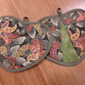 May include: Two heart-shaped pot holders with a floral pattern in red, yellow, and blue on a dark background. The edges are trimmed with a green border. One has a green fabric accent.