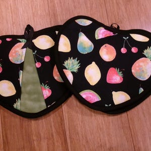 May include: Two heart-shaped pot holders with a black background and a colorful fruit pattern. The design includes lemons, strawberries, cherries, pineapples, pears, and watermelon slices. Each pot holder has a black trim and a loop for hanging.