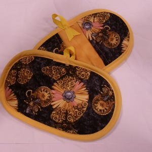K&ouml;nnte beinhalten: Zwei ovale Topflappen mit einem schwarzen Blumen- und Uhrendesign. Sie haben einen gelben Rand und eine Aufh&auml;ngeschlaufe. Das Design enth&auml;lt rosa, gelbe und blaue Details. Ma&szlig;e in Zentimetern.