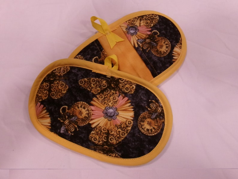 K&ouml;nnte beinhalten: Zwei ovale Topflappen mit einem schwarzen Blumen- und Uhrendesign. Sie haben einen gelben Rand und eine Aufh&auml;ngeschlaufe. Das Design enth&auml;lt rosa, gelbe und blaue Details. Ma&szlig;e in Zentimetern.