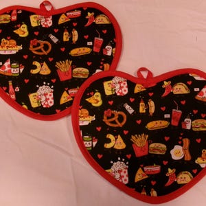 May include: Two heart-shaped pot holders with a black background and a red trim. The fabric is patterned with various food illustrations, including pizza, fries, and tacos. Each pot holder has a red loop for hanging.